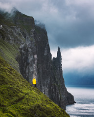 Wall Mural - Tourist in yellow jacket looks at Witches Finger cliffs from Trollkonufingur viewpoint. Vagar island, Faroe Islands, Denmark.