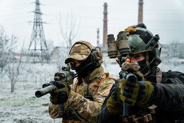 Wall Mural - Two men in camouflage cloth and black uniform with machineguns with factory on background. Soldiers with muchinegun aims aiming standing beside wall