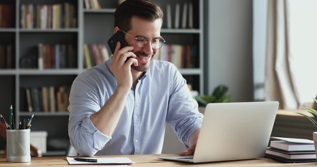 smiling business man using laptop talking on cell phone sits at desk. happy confident male professio