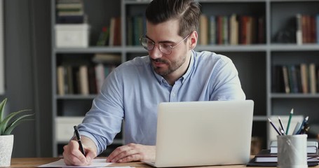 Focused business man working on laptop computer making notes sitting at home office desk. Young adult male professional manager doing marketing research writing checklist on paper at workplace.