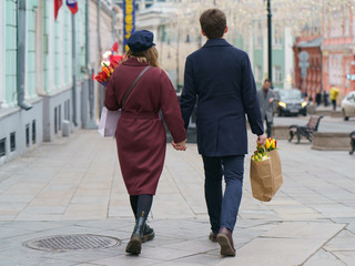 Festive mood. People on spring city street. Holidays concepts. Portrait of couple of young man and woman with bouquet of spring flowers (red, yellow tulips). Back view