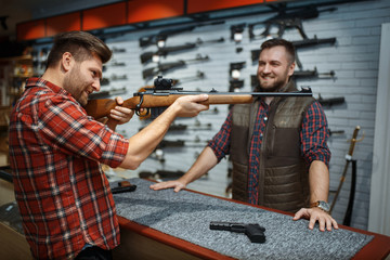 Man aims with rifle, seller at counter in gun shop