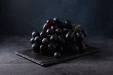 A Cluster Of Juicy Black Grapes Lies On A Black Slate On A Dark Gray Table