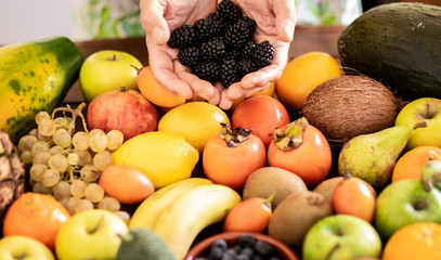 Two human hands holding some blackberries and background with different kind of fresh fruits.  Healthy eating concept and detox diet. Vegan and vegetarian food.