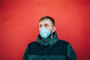 young man with a beard is standing in a medical mask that protects against coronovirus and other diseases and viruses against a bright red wall