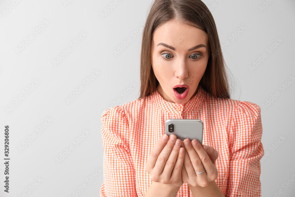 Surprised young woman with mobile phone on light background