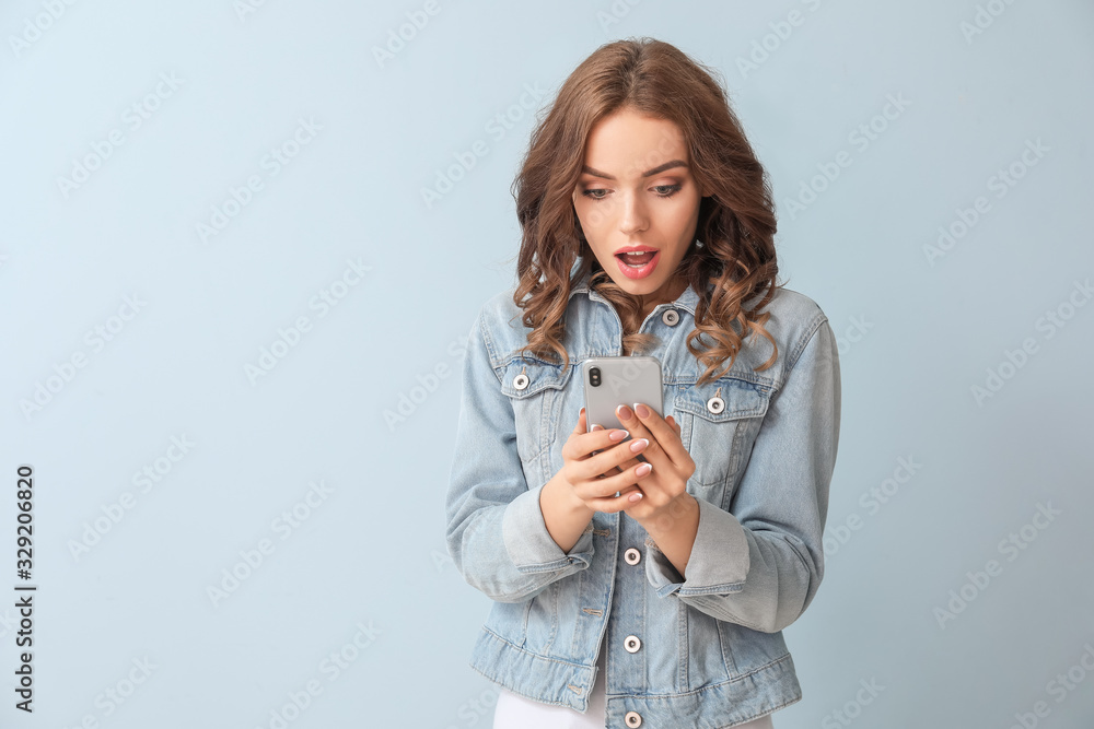 Surprised young woman with mobile phone on color background