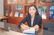 © Have a nice day  - asian woman hotel receptionist standing behind a counter with pen and a notebook next to a computer monitor smiling, wearing a suit and white shirt with wooden cabinets and books at the background.