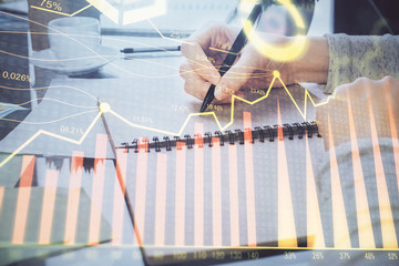 Hand taking notes in notepad. Forex chart holograms in front. Concept of research. Double exposure