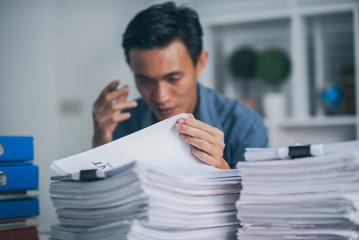 Young business man doing financial reports feeling stress with stack of papers.