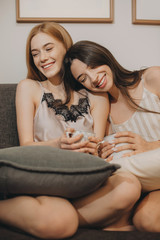 ginger caucasian girl sitting on the sofa near her brunette sister while drinking a coffee and havin