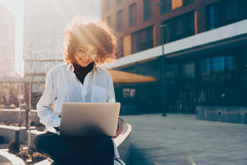 smiling curly haired caucasian woman working outside on her computer while sitting on stone stairs a