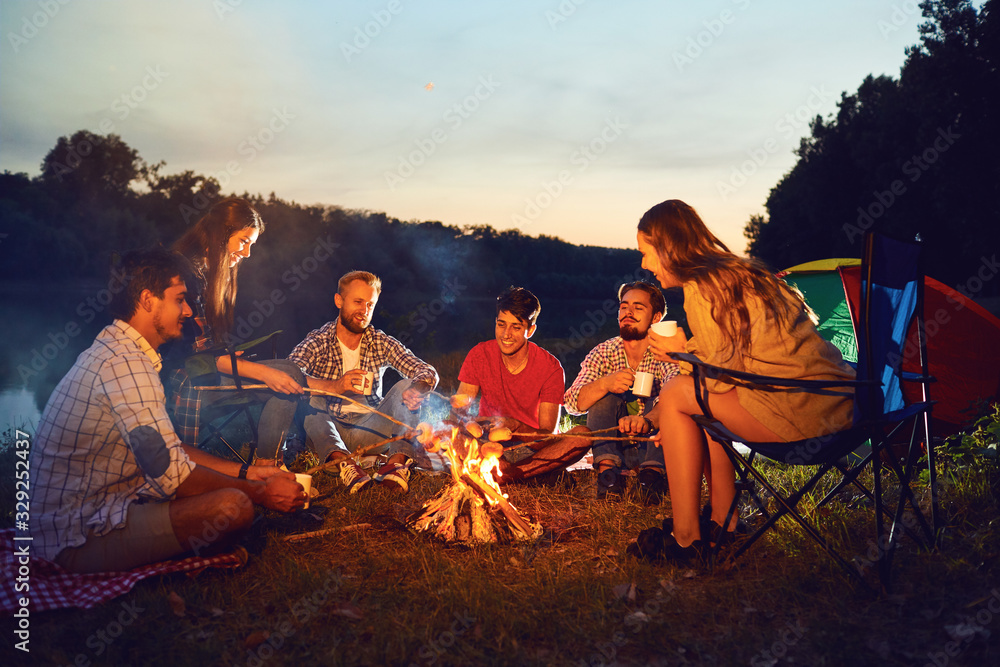 Friends are laughing sitting by the fire next to the tent. - obrazy, fototapety, plakaty 