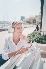 Wall Mural - Enjoying the sound. Happy young woman with earphones listening music on smartphone while sitting on city embankment sea promenade.