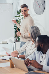 Wall Mural - multicultural colleagues sitting at table and looking at smiling businessman during presentation