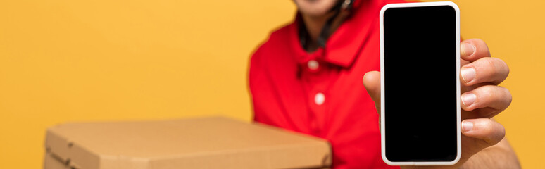 Wall Mural - cropped view of happy delivery man in red uniform holding pizza boxes and smartphone with blank screen isolated on yellow, panoramic shot