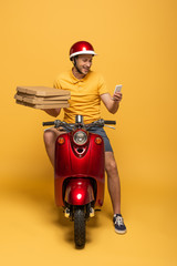 Wall Mural - smiling delivery man in yellow uniform riding scooter with pizza boxes and using smartphone on yellow background