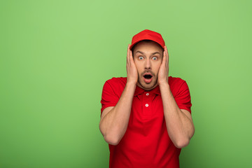 Wall Mural - scared delivery man in red uniform with open mouth touching face on green