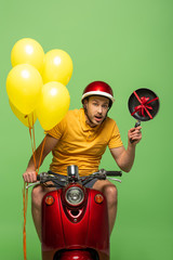 Wall Mural - confused delivery man in yellow uniform on scooter with frying pan and balloons isolated on green