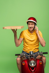 Wall Mural - smiling delivery man in yellow uniform on scooter with pizza talking on smartphone isolated on green