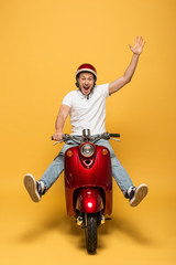 Wall Mural - happy delivery man in helmet riding scooter with raised hand on yellow background