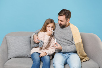 Wall Mural - Man in knitted sweater with sick child baby girl. Father little daughter isolated on blue background. Love family parenthood childhood concept. Sit on couch, having runny nose, blowing nose to napkin.