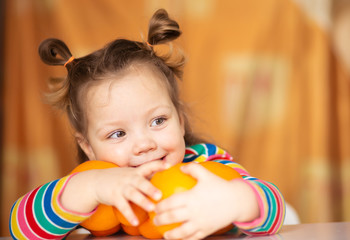 Little girl, 3 year old baby, with a ponytail hairstyle in a colorful colorful striped jacket hugs a lot of appelsins