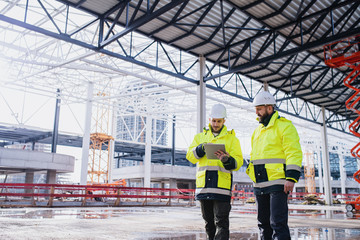 Men engineers standing outdoors on construction site, using tablet.