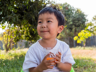 Asian little child boy holding glass of orange juice with smiling happy face outdoor nature background at home.  Vitamin C nutrition for healthy concept.