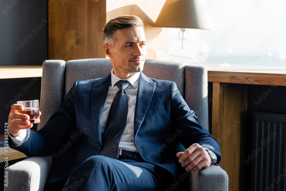 smiling businessman in suit sitting in armchair and holding glass - obrazy, fototapety, plakaty 