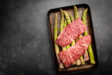 Wall Mural - Raw Steak with green asparagus on tray on black background, top view