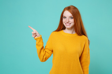 Smiling young redhead woman girl in yellow sweater posing isolated on blue turquoise background studio portrait. People emotions lifestyle concept. Mock up copy space. Pointing index finger aside up.