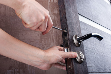 Handyman with a screwdriver installs and adjusts steel door lock in a wooden casement. Locksmith repairs and replaces locking mechanism in the living room. Installation and maintenance the doors.