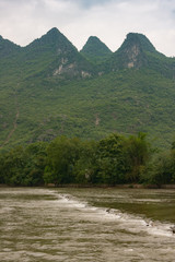 Guilin, China - May 10, 2010: Along Li River. Portrait of chain of green forested karst mountains under blue cloudscape with white line of rapid in greenish water. Green tree belt.