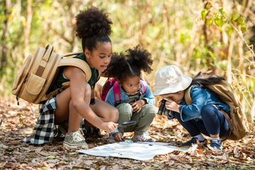 Group of happy pretty little girl hiking together with backpacks and sitting on forest dirt road with looking at the map for exploring the forest. Three kids having fun adventuring in sunny summer day