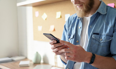 Close up view of smiling man professional user standing at home office holding smart phone in hands text message. Young student using cellphone application technology modern gadget concept. Copyspace.