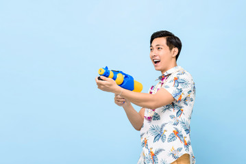 Smiling handsome Asian man playing with water gun isolated on studio blue background for Songkran festival in Thailand and southeast Asia