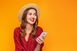 © Evgenia - Portrait of a young woman in a red summer dress, a hat with red lipstick holds a mobile phone, cell in her hands, laughs, smiles and looks at copy space isolated on a yellow, orange background