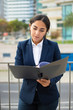 © Mangostar - Focused businesswoman reading papers. Front view of concentrated young businesswoman in formal wear holding folder with documents on street. Paperwork concept