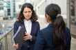 © Mangostar - Serious businesswomen discussing work. Professional female colleagues in formal wear standing on urban city street and discussing papers. Cooperation concept