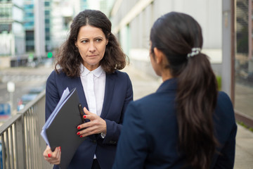 Serious businesswomen discussing work. Professional female colleagues in formal wear standing on urban city street and discussing papers. Cooperation concept