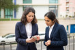 © Mangostar - Focused businesswomen using smartphone. Female colleagues in formal wear standing on street and using cell phone together. Business and technology concept