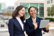 © Mangostar - Businesswomen having video chat outdoors. Cheerful female colleagues standing on city street and talking via tablet pc. Communication concept