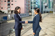 © Mangostar - Content businesswomen shaking hands. Side view of cheerful female colleagues in formal wear standing on street and greeting each other. Partnership concept