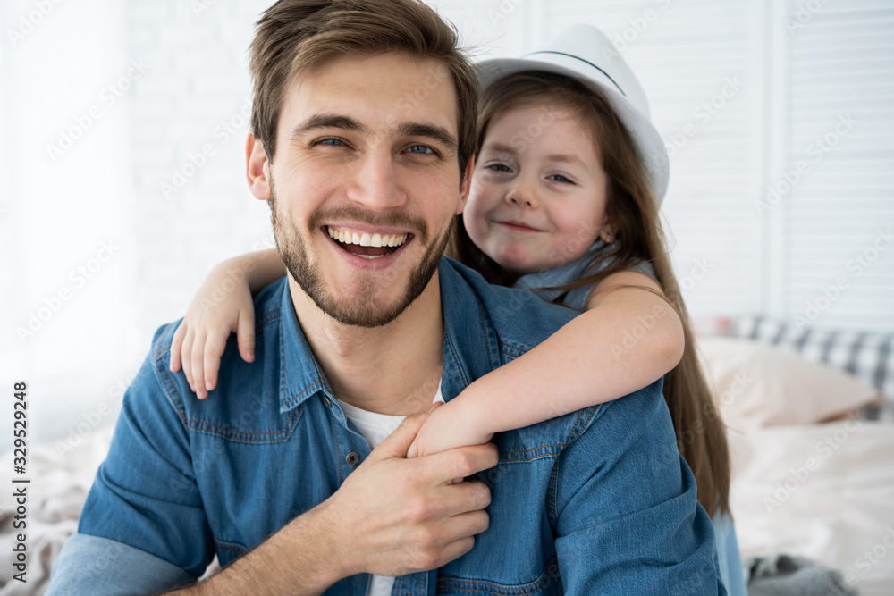 Portrait of handsome father and his cute daughter hugging, looking at camera and smiling while sitting on sofa at home. - obrazy, fototapety, plakaty 