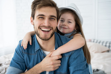 Wall Mural - Portrait of handsome father and his cute daughter hugging, looking at camera and smiling while sitting on sofa at home.