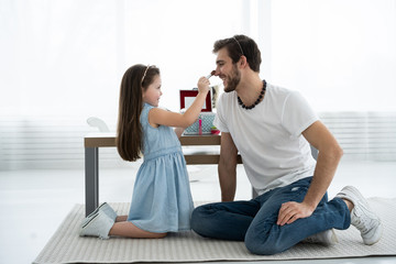 Wall Mural - Cute little daughter and her handsome young dad in crowns are playing together in child's room. Girl is doing her dad a makeup.