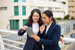 © Mangostar - Content businesswomen using tablet pc. Cheerful focused female colleagues standing with digital tablet on urban city street. Wireless technology concept