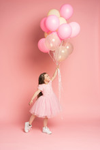 Happy Celebration Of Birthday Party With Flying Balloons Of Charming Cute Little Girl In Tulle Dress Smiling To Camera Isolated On Pink Background. Charming Smile, Expressing Happiness