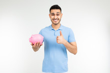 Brutal Man In A Blue Shirt Holds A Piggy Bank And Shows A Super Class On A White Background With Copy Space